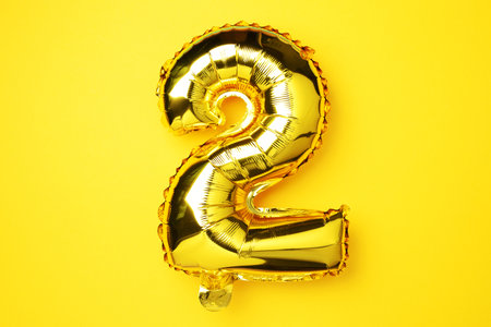 Creative layout. Golden foil balloon number and digit two 2. Birthday greeting card. Anniversary concept. Top view. Copy space. Stylish gold numeral over yellow background. Numerical digit.の写真素材