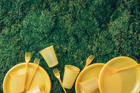 Environmental pollution. Disposable plastic tableware - plastic plates, forks, spoons, knives. Yellow plastic tableware on green grass, moss background. Top view. Copy space. Ban single use plasticの写真素材