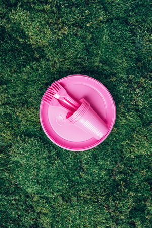 Environmental pollution. Disposable plastic tableware - plastic plates, forks, spoons, knives. Pink plastic tableware on green grass, moss background. Top view. Copy space. Ban single use plasticの写真素材