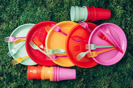 Disposable plastic tableware - plates, forks, spoons. Colored plastic disposable tableware on green grass moss background. Top view. Copy space. Birthday picnic utensil. Recycling plastic and ecologyの写真素材