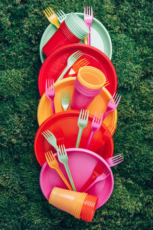 Colored plastic disposable tableware on green grass moss background. Top view. Copy space. Birthday picnic utensil. Recycling plastic and ecology. Disposable plastic tableware - plates, forks, spoonsの写真素材