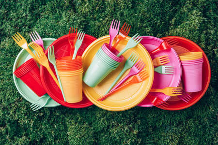 Disposable plastic tableware - plates, forks, spoons. Colored plastic disposable tableware on green grass moss background. Top view. Copy space. Birthday picnic utensil. Recycling plastic and ecologyの写真素材