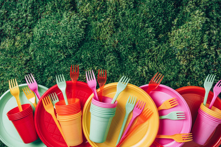 Colored plastic disposable tableware on green grass moss background. Top view. Copy space. Birthday picnic utensil. Recycling plastic and ecology. Disposable plastic tableware - plates, forks, spoonsの写真素材