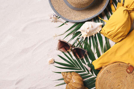 Female beach accessories: swimsuit, bikini, rattan bag, straw hat, shells, sunglasses, palm leaves on sand background. Exotic, tropical mood. Summer vacation, travel concept. Flat lay. Copy space.の写真素材