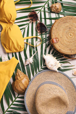 Womans beach accessories: swimsuit, bikini, rattan bag, straw hat, shells, sunglasses, palm leaves on sand background. Exotic, tropical mood. Summer vacation, travel concept. Flat lay. Copy space.の写真素材