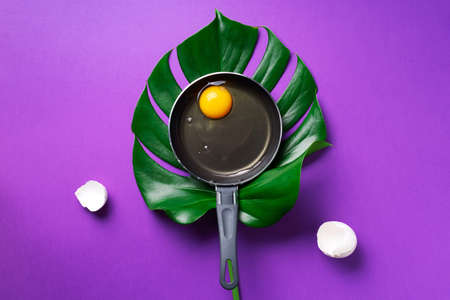 Organic egg in pan over monstera leaf on violet background. Minimalistic food concept. Top view. Creative layoutの写真素材