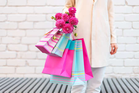 Pink flowers in female hands and shopping bags on brick background. Birthday, Mothers, Womens, Wedding Day concept. Bunch of paper gift bagsの写真素材