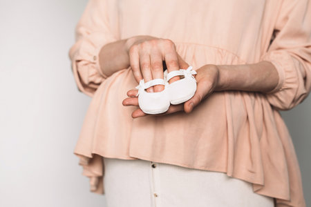 Pregnant woman in pink dress holding small shoes for unborn baby on belly, preparing go to maternity hospital for childbirth. Pregnancy, maternity, preparation, baby expectation concept. Copy spaceの写真素材