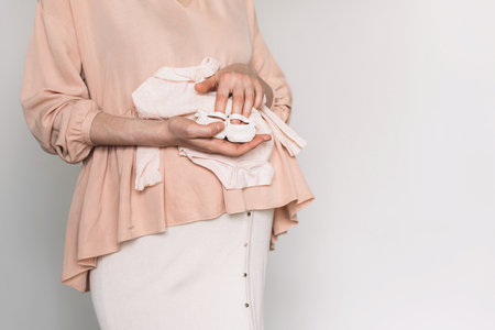 Pregnant woman in pink dress holding small shoes for unborn baby on belly, preparing go to maternity hospital for childbirth. Pregnancy, maternity, preparation, baby expectation concept. Copy spaceの写真素材