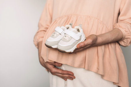 Pregnancy, maternity, preparation, baby expectation concept. Pregnant woman in pink dress holding small shoes for unborn baby on belly, preparing go to maternity hospital for childbirthの写真素材