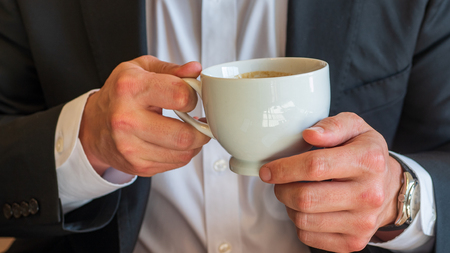 Man holding a cup of coffee with cream in white ceramic cup, dressed in white dress shirt and black business suit and wearing a watchの写真素材