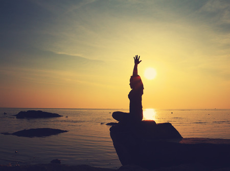 Silhouette yoga girl by the beach at sunrise doing meditation on rock  in vintage styleの写真素材