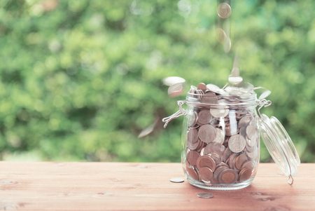 Money coins falling in glass jar on old  wooden table with green nature background,saving money conceptの写真素材