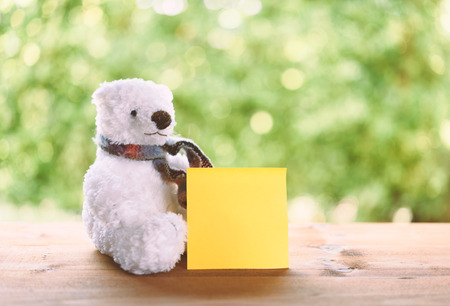 White puffy teddy bear toy sit with yellow sticky note on wood table with green blurred backgroundの写真素材