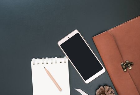 Workspace with mobile phone, leather bag notebook, pen, pencil and stainless steel cutter on dark background, top viewの写真素材