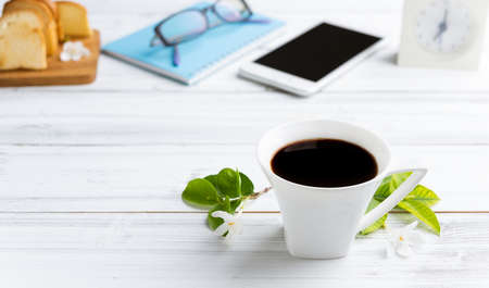 Breakfast set with coffee cup,crispy butter bread,mobile phone,eye glasses,and blue notebook on white wood tableの写真素材