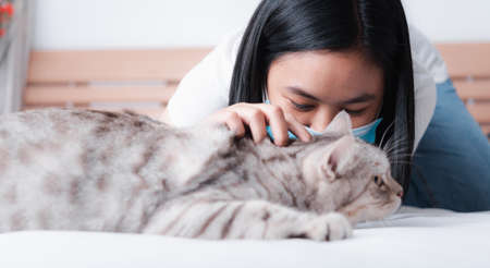 Beautiful smiling woman face wearing face mask to protect from virus or illness and play with her tabby cat on  white bedの写真素材