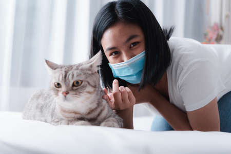Beautiful smiling woman face wearing face mask to protect from virus or illness and play with her tabby cat on  white bedの写真素材