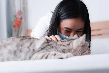 Beautiful smiling woman face wearing face mask to protect from virus or illness and play with her tabby cat on  white bedの写真素材