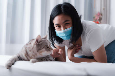 Beautiful smiling woman face wearing face mask to protect from virus or illness and play with her tabby cat on  white bedの写真素材