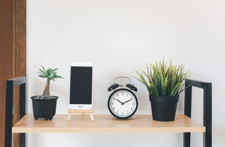Wooden shelf with mobile phone,grass in pot, greenery in vase, alarm  and black board  over white wall  decoration in living room at homeの写真素材
