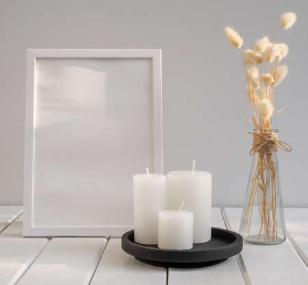 Mock up white poster frame,candles  and beautiful Lagurus ovatus dried flowers in modern glass vase  composition over white wooden table room interiorの写真素材