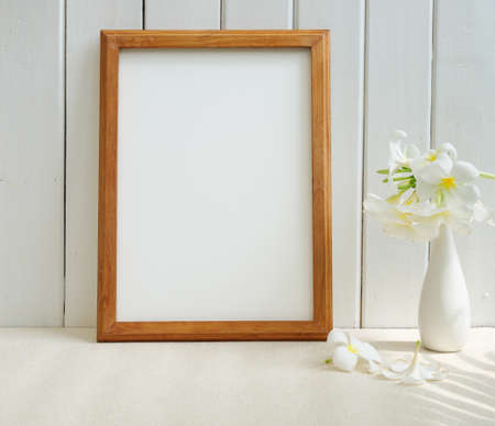 Mock up poster wooden frame and beautiful Frangipani tropical flower in modern white vase on beige table and wood wall background with palm leaf shadowの写真素材