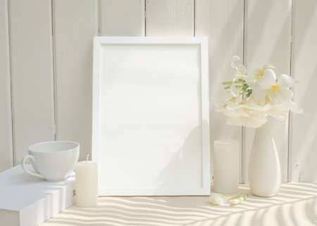 Blank mockup picture frame,coffee cup candle,and Plumeria flower in ceramic white vase on beige table with white wood background,long palm shadowの写真素材