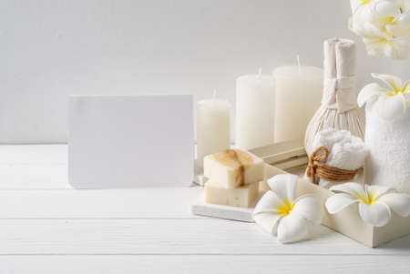 Spa card invitation with massage  herbal ball,plumber flower in vase,coconut coffee soap,white towels and candle on white wood table background,soft white tone still lifeの写真素材