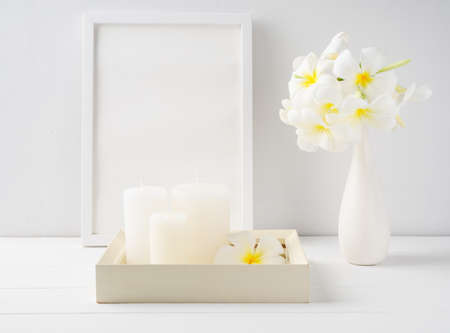 White candles on white wood  tray,mock up poster frame and Plumeria flowers in modern ceramic vase  over white wooden table room interior,spa soft tone still lifeの写真素材
