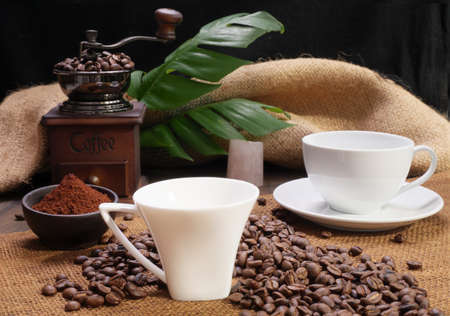 Two coffee cups,ground coffee ,roasted coffee beans,grinder, and monstera leave on wooden table with burlap  backgroundの写真素材