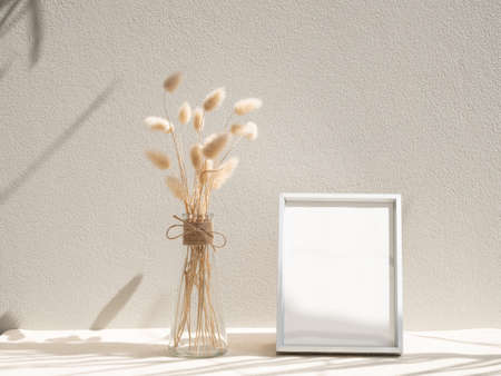 Blank mockup picture frame and dried flower in glass vase on beige table and cement wall backgroundの写真素材