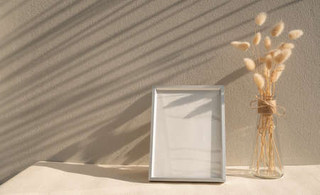 Blank mockup picture frame and dried flower in glass vase on beige table and cement wall backgroundの写真素材