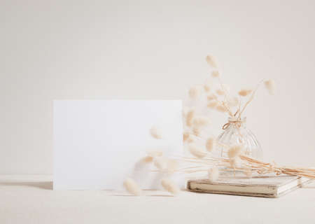 Blank white greeting card mock up.decoration with dried Lagurus ovatus flowers composition in modern glass vase and craft book  on  beige table and cement wall backgroundの写真素材