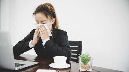 Sick young asian business woman sitting alone at work sneeze holding tissue handkerchief and blowing wiping her running nose.の写真素材