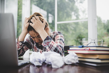 Young frustrated woman working at office desk in front of laptop suffering from chronic daily headaches, treatment online, appointing to a medical consultation, electromagnetic radiation, sick payの写真素材