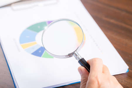 professional business woman hand holding and using magnifying glass search finance document paper on desk in office, businesswoman looking at data with analyzing statisticsの写真素材