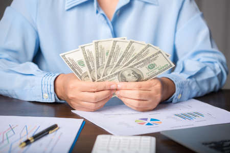 Close up young business woman using counting cash money one hundred dollar bills, checking financial documents, sitting at table with papers and tax form, managing planning budget, accounting expensesの写真素材