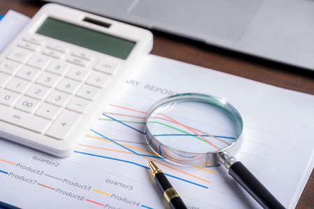 magnifying glass with business document financial paper chart statistic report on desk in office, closeup calculating machine with paperwork on table concept for analysis, market, bankingの写真素材