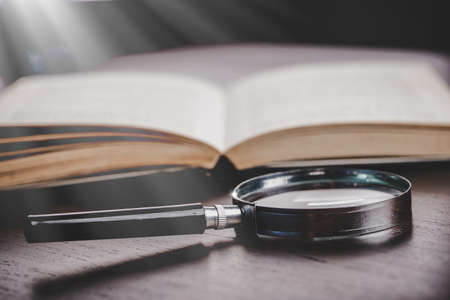 open book with magnifying glass on wooden desk in information library of school or university,...