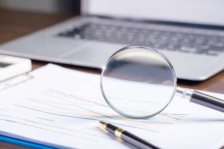 magnifying glass with business document financial paper chart statistic report on desk in office, closeup calculating machine with paperwork on table concept for analysis, market, bankingの写真素材