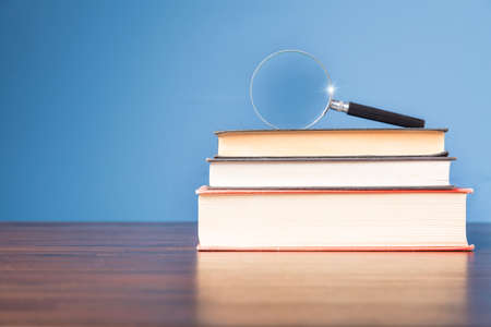 Stack book with magnifying glass on wooden desk in information library of school or university, concept for education, reading, study, copy space on blue background.の写真素材