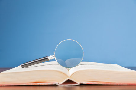 open book with magnifying glass on wooden desk in information library of school or university, concept for education,reading , study, copy space and blue background.の写真素材