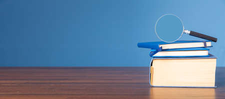 Stack book with magnifying glass on wooden desk in information library of school or university, concept for education, reading, study, copy space on blue background.の写真素材