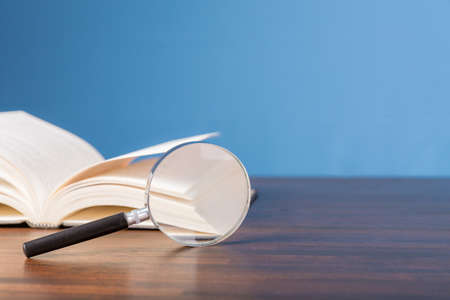 open book with magnifying glass on wooden desk in information library of school or university, concept for education,reading , study, copy space and blue background.の写真素材