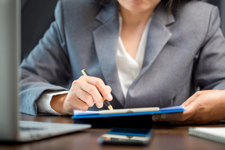 Workplace closeup person professional businesswoman sitting at desk hold pen signing or signature contract paper. Employee woman writing agreement document on paperwork form corporate at work officeの写真素材