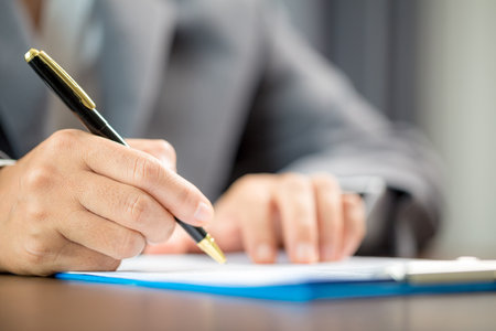 Workplace closeup person professional businesswoman sitting at desk hold pen signing or signature contract paper. Employee woman writing agreement document on paperwork form corporate at work officeの写真素材