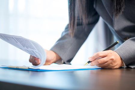 Workplace closeup person professional businesswoman sitting at desk hold pen signing or signature contract paper. Employee woman writing agreement document on paperwork form corporate at work officeの写真素材