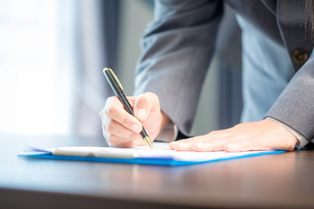 Workplace closeup person professional businesswoman sitting at desk hold pen signing or signature contract paper. Employee woman writing agreement document on paperwork form corporate at work officeの写真素材