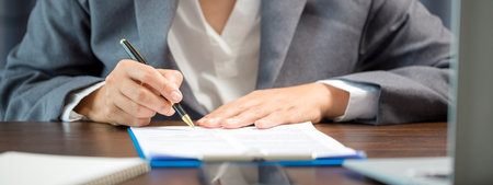 Workplace closeup person professional businesswoman sitting at desk hold pen signing or signature contract paper. Employee woman writing agreement document on paperwork form corporate at work officeの写真素材